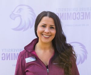 Smiling woman with long brown hair wearing a maroon jacket with a name tag, standing in front of a backdrop with a faint eagle logo and partial text.