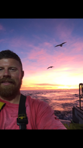 Jason Kusiak smiles for a selfie in front of a sunset over the ocean. Two birds soar overhead.