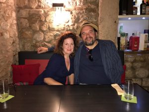 Two people sitting together at a table in a rustic restaurant, with a stone wall and light fixture behind them. Two empty glasses on green coasters and a folded napkin are on the table.