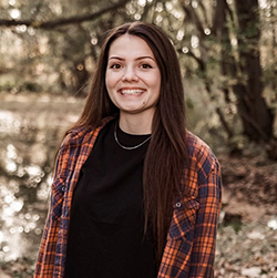 A smiling young woman in a plaid shirt stands by a serene pond, surrounded by greenery.