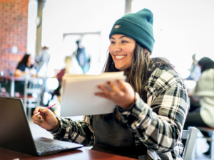 Young woman in a plaid jacket and beanie working on a laptop, holding a notebook and pen, in a bright, indoor setting with other people in the background.