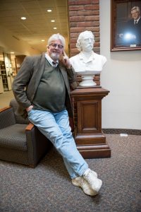 Phil Adamo perches on the arm of a chair, resting his elbow on a plinth displaying a bust in the Lindell Library