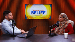 Adam Duxter and Najeeba Syeed smile, facing each other at a table with microphones