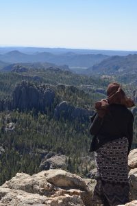 Female student standing on top of rocks at the peak of Black Elk Peak looking over the Black Hills. She is wearing a black and white shirt, black sweater and maroon hijab.