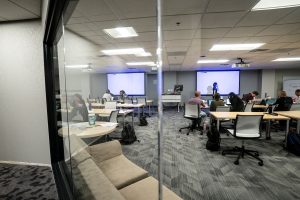 academic lecture room with rectangular desks, two projection screens in background, couch in foreground