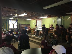 Students sit around tables listening as a woman talks.