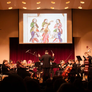 Orchestra performing on stage with a conductor, screen above showing a historical illustration of musicians.