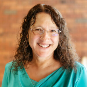 Person with curly hair and glasses, smiling in front of a blurred brick wall.