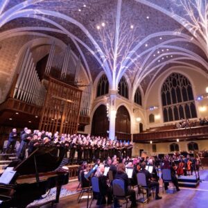 A choir and orchestra performing in a grand concert hall with a vaulted, illuminated ceiling.