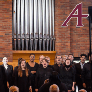 A choir group singing in front of a pipe organ in a brick-walled venue, with a stylized letter "A" in the top right corner.