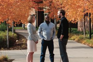 three people conversing on a sidewalk surrounded by fall foliage