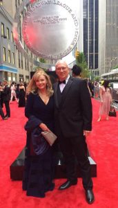 Two people in formal attire on a red carpet in front of a metallic circular sculpture with text.