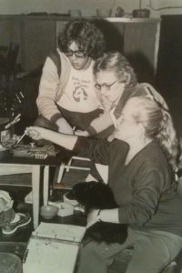 Three people working at a table with small tools and a dog on one person's lap.