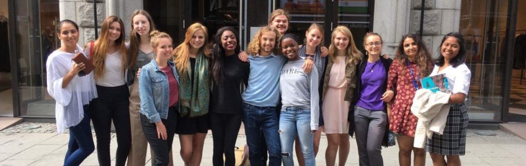  A group of thirteen people standing outside a building with glass doors, smiling and with arms around each other.