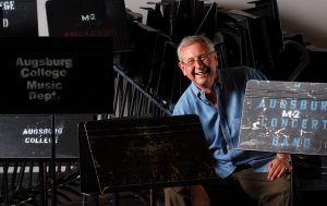 Older man in a blue shirt smiling among stacked black music stands.
