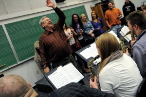 A man enthusiastically gestures while leading a music group in a classroom setting.