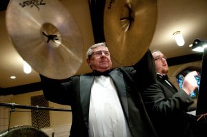  A man in a tuxedo holding a pair of raised cymbals, with another man in a tuxedo in the background.