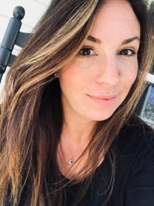 Close-up of a woman with long brown hair and lighter streaks, wearing a black top and a delicate necklace, and smiling slightly.