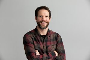 Man with a beard, wearing a plaid shirt, standing with arms crossed against a gray background.