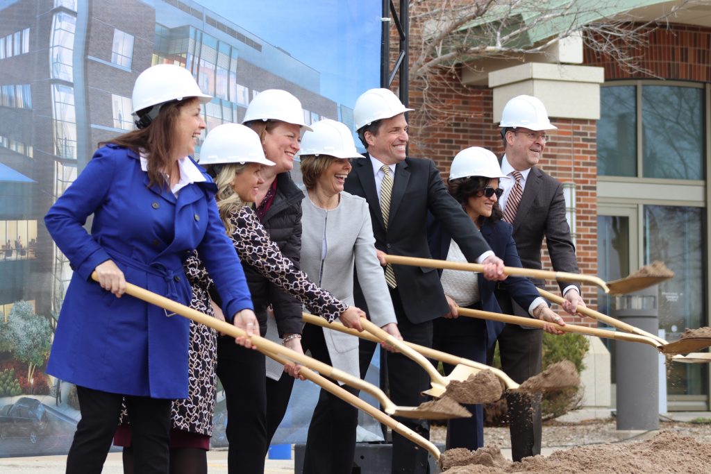 A photo of the group in hard hats holding shovels with dirt. From Left to right,: Jill Billings, Sarah Erkkinen, Amy Alkire, Nancy Mueller, Paul Mueller, Lisa Archer, and Matt Entenza