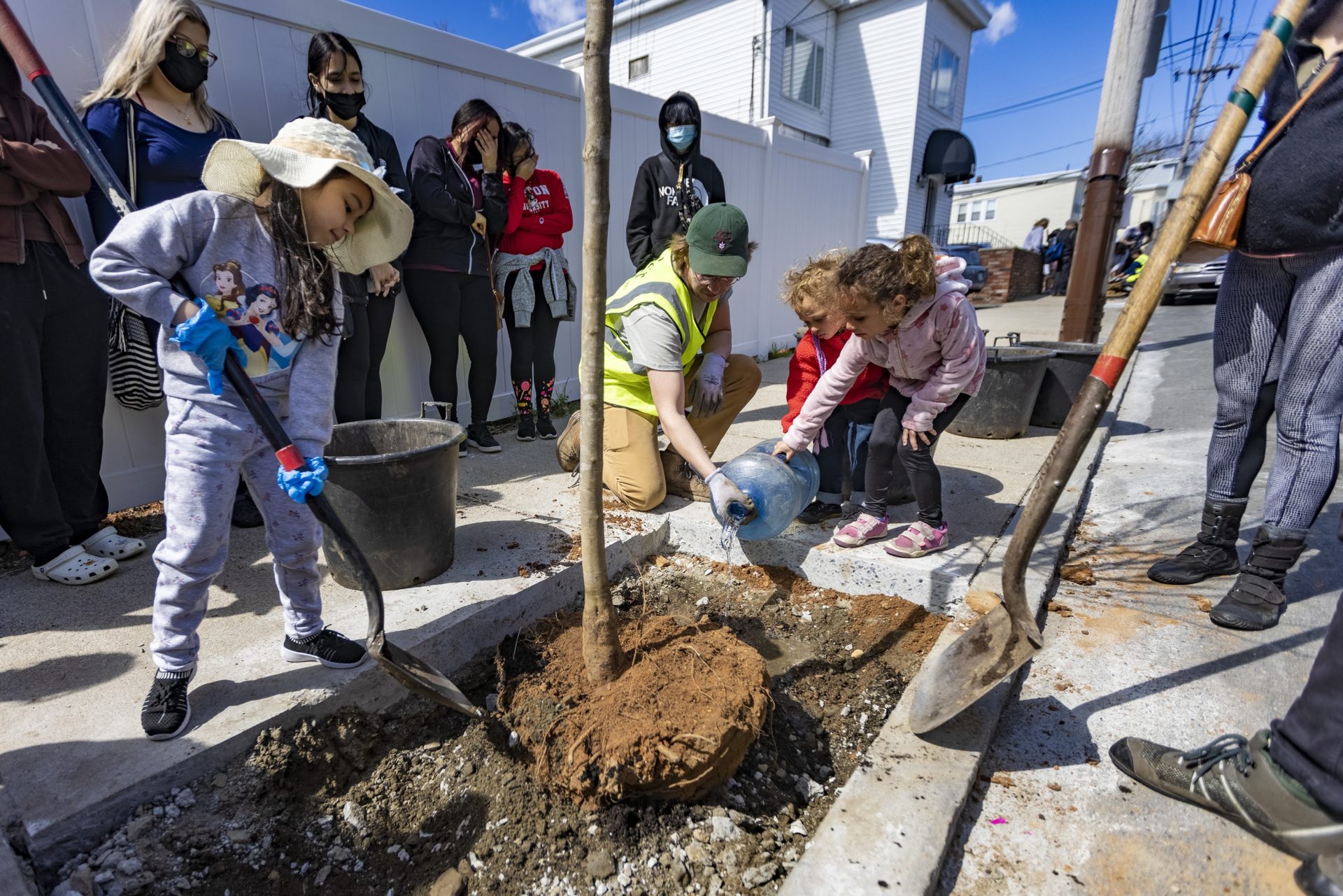 five-year-old girl shovels dirt as man helps a two-year-old boy and four-year-old girl water the root of a tree in the ground, a crowd gathers around them