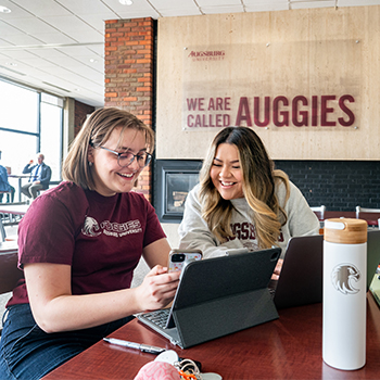 Two young women smiling and looking at a tablet in a university lounge area.