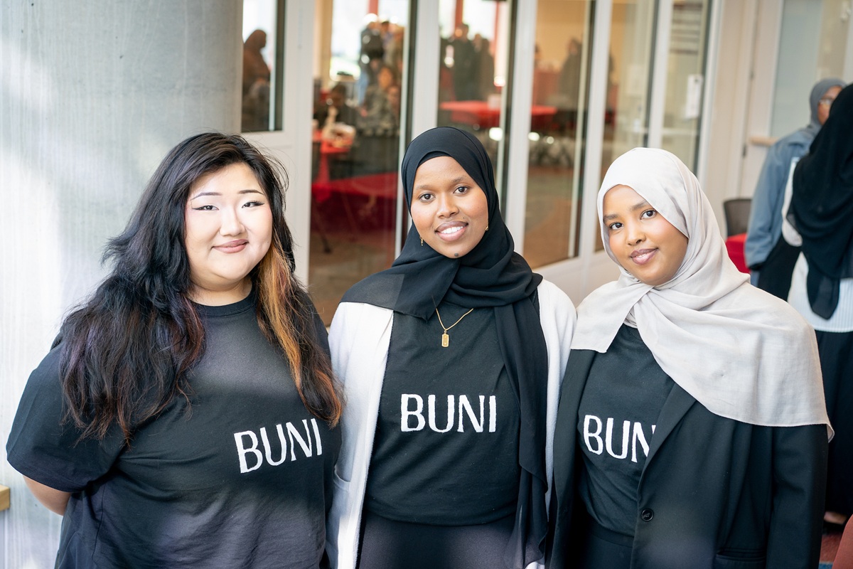 Ngawang Nima, Hodo Abdi, and Rahma Mohamed stand side by side indoors, smiling at the camera, each wearing a black “BUNI” T-shirt; two wear headscarves and one has long dark hair.