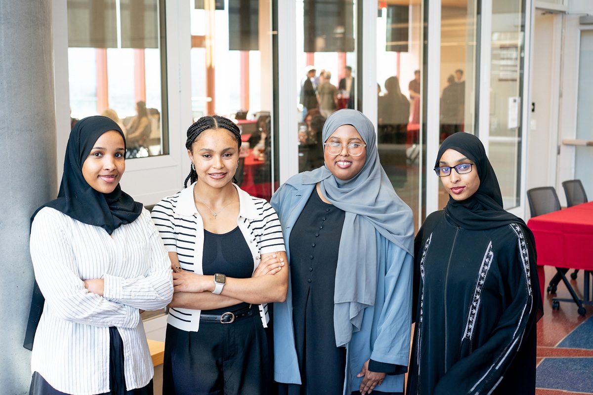Sena Dangul, Samia Ahmed, Hodan Huran, and Sumeya Mahamed stand together indoors, smiling at the camera, with a glass-walled room and people in the background.