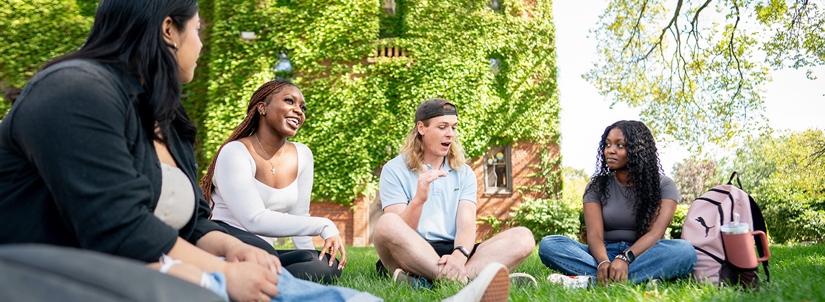 A group of four students relaxes on the grass in front of Memorial Hall at Augsburg University.
