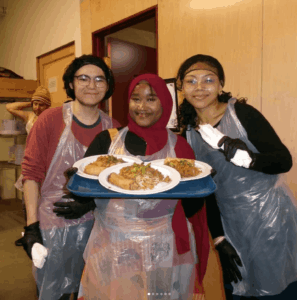 A student holds a plastic tray with 3 plates of food while two students pose at each side. A service project by the participants of the Alternative Spring Break trip to New York City.