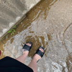 Individual stepping in water that is flowing by the side of a half-wall by the riverside. 