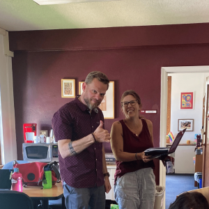 Jeremy Myers and Kristina Fruge in the CCV office. Jeremy is giving a thumbs up and Kristina is smiling with a laptop in her arms. 