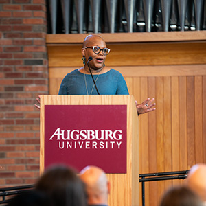 Person speaking at a podium with "Augsburg University" text and a background of brick and wood paneling.