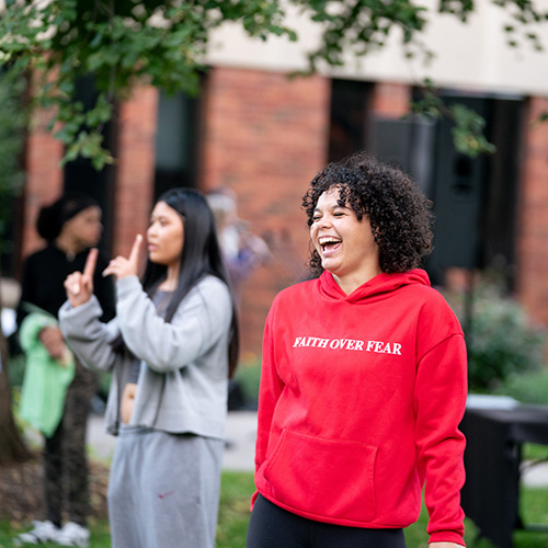 A girl wearing a bright red shirt that says "Faith Over Fear" laughing outside of Foss Center.