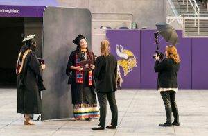 Graduates stand in front of photo backdrop and professional photographers in U.S. Bank Stadium