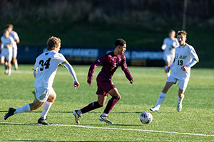 Soccer player in maroon dribbling the ball between two opponents in white jerseys on a sunlit field.