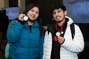 Two people standing together, each holding a medal, with a screen in the background showing the text "Auggie Entrepreneur".