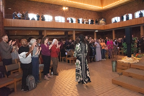 2025 Interfaith Symposium A large room with people standing and applauding in front of a person in a patterned robe.