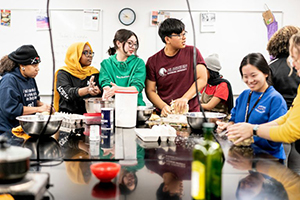 A diverse group of people gathered around a kitchen counter preparing food in a classroom setting.