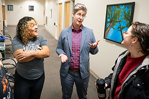 Three people talking in a hallway with a painting on the wall.