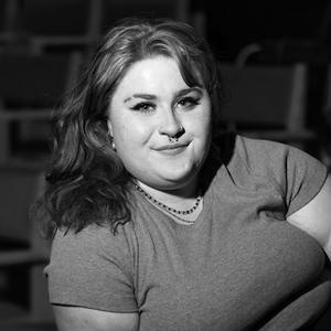 Black-and-white photo of a young person with wavy hair and a septum piercing, seated in front of blurred rows of seats.