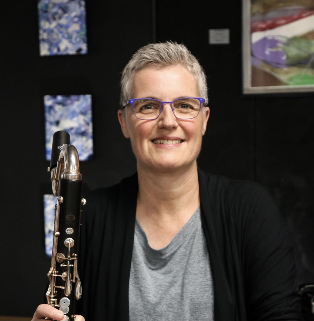 A person with short gray hair in blue glasses holding a clarinet, with abstract paintings in the background.