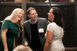 A woman in a green dress, man in a suit, and a woman in a silver dress smile at each other as they converse during an event.