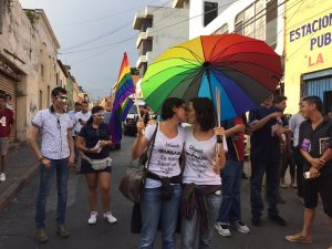 two people kiss in a parade under a rainbow umbrella