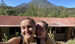 Two young women smile for a selfie in front of a mountainous background