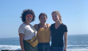 three women stand on a large rock with the blue ocean behind them