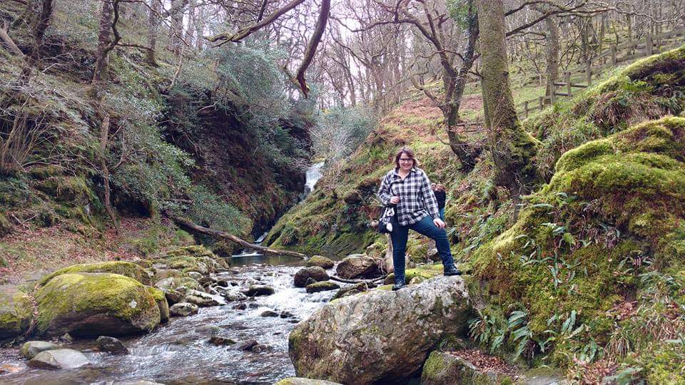 a young woman stands at a creek with green forest scenery behind her