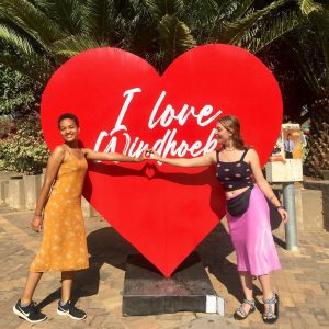 Two students stand with their hands in the heart shape in front of a heart sign that reads I Love Windhoek