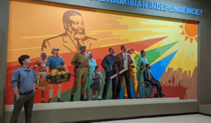 A student stands in front of a painted mural of several people under the Namibia flag