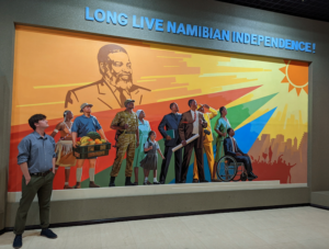 A student stands in front of a painted mural of several people under the Namibia flag